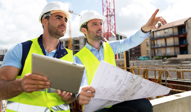Two men in hard hats holding a tablet and blue prints, pointing off camera